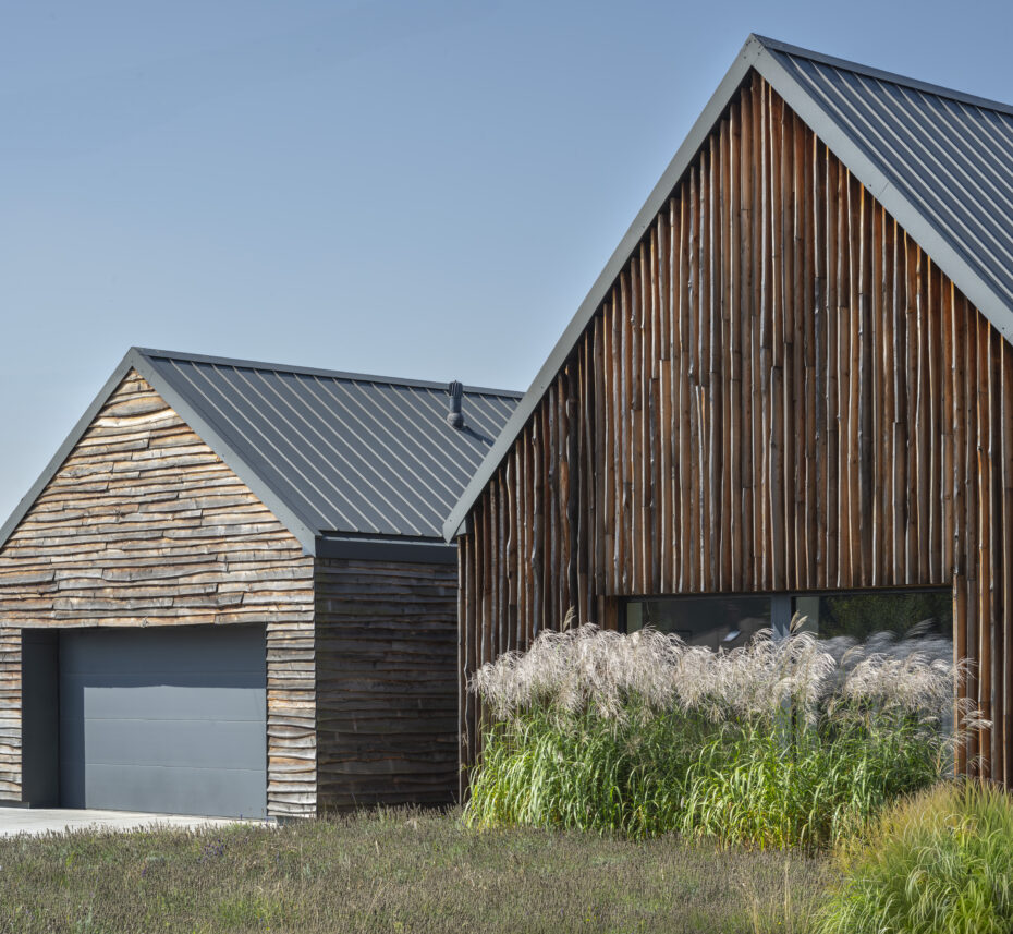 Widok z ulicy na dom z drewnianą nieheblowaną elewacją z wysoką trawą w przedpolu.