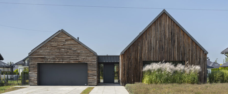 Widok z ulicy na podjazd i dom z drewnianą nieheblowaną elewacją z wysoką trawą w przedpolu