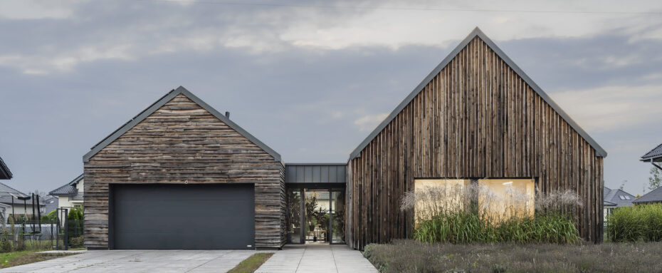 Widok z ulicy na podjazd i dom z drewnianą nieheblowaną elewacją z wysoką trawą w przedpolu